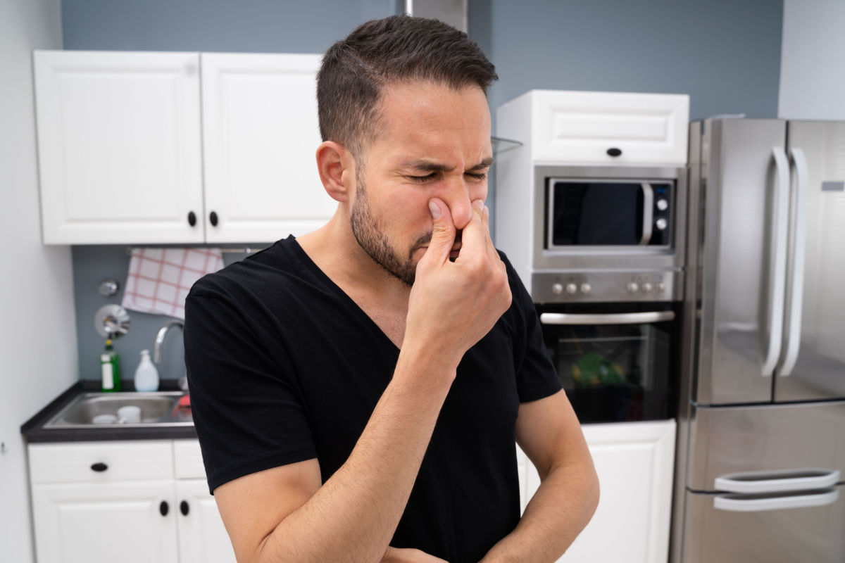 A man in a kitchen pinching his nose due to a bad smell, indicating an unpleasant odor, possibly from a plumbing or drainage issue.